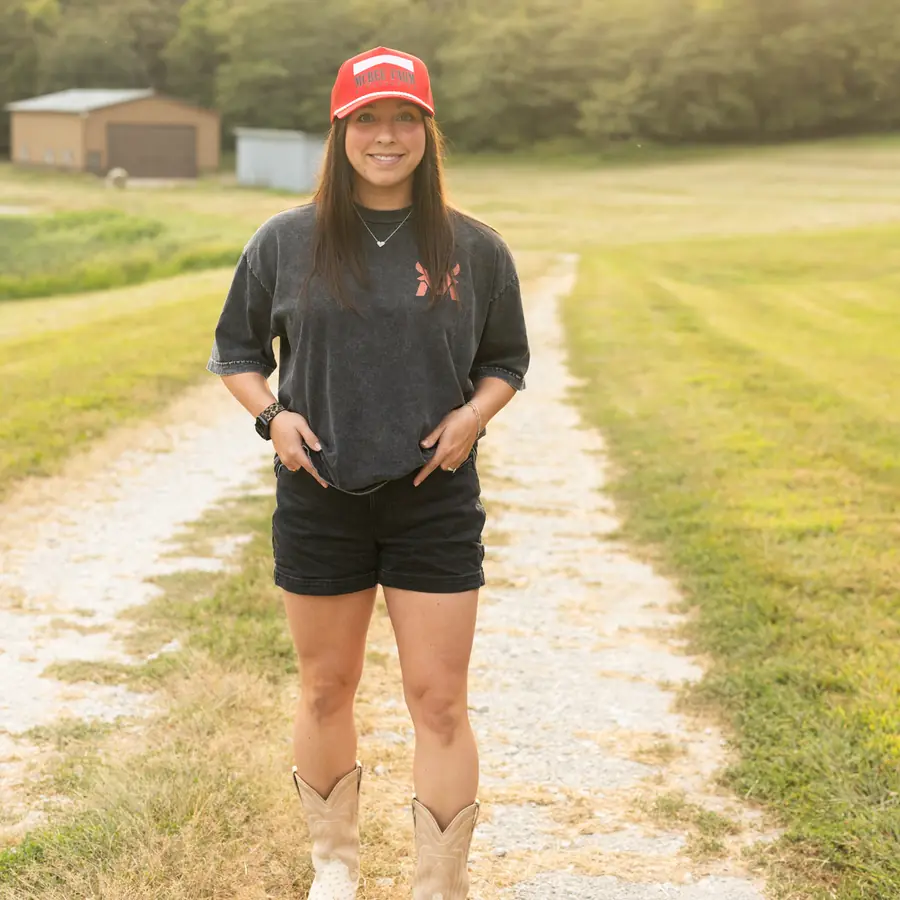 Alli McBee in a red McBee Farm trucker hat and cowboy boots on a gravel farm road