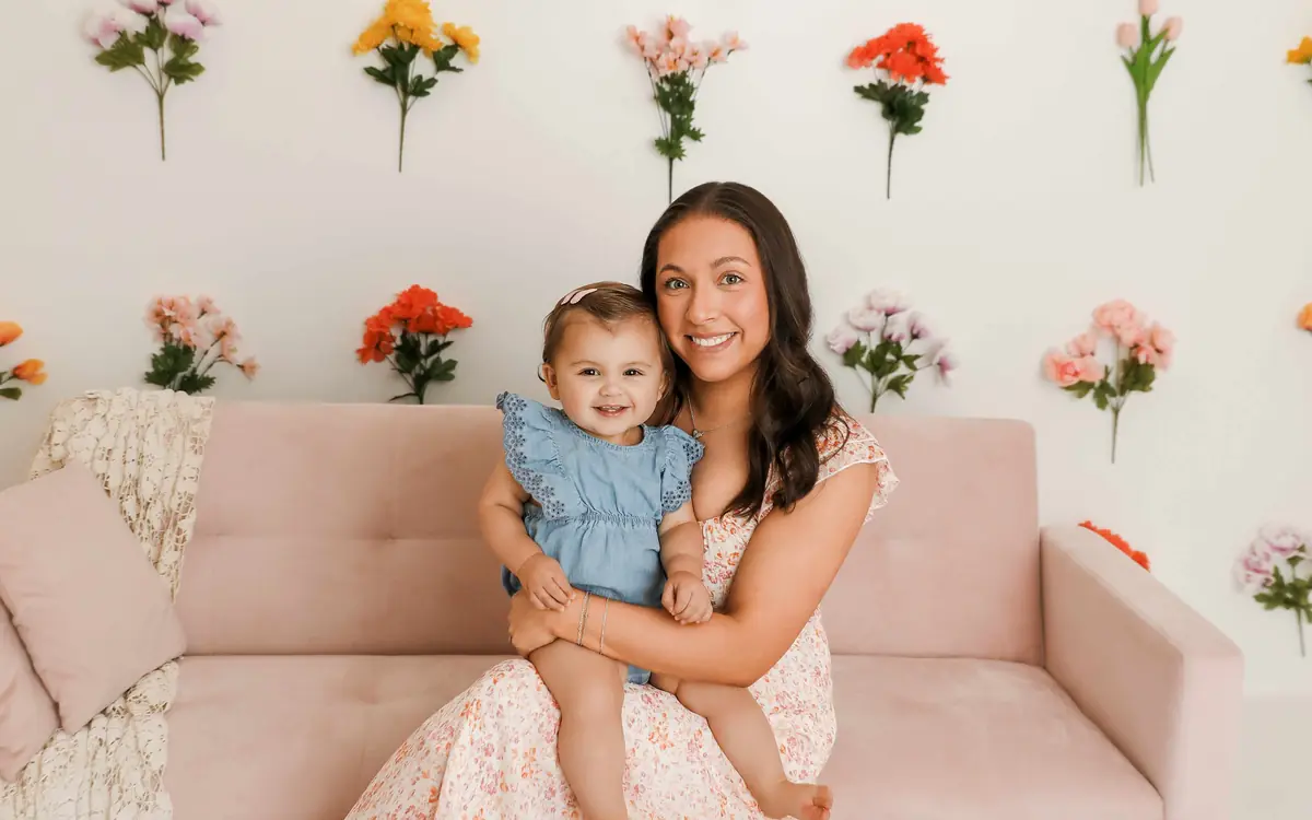 Alli McBee holding Summer on a pink couch in front of a wall of flowers